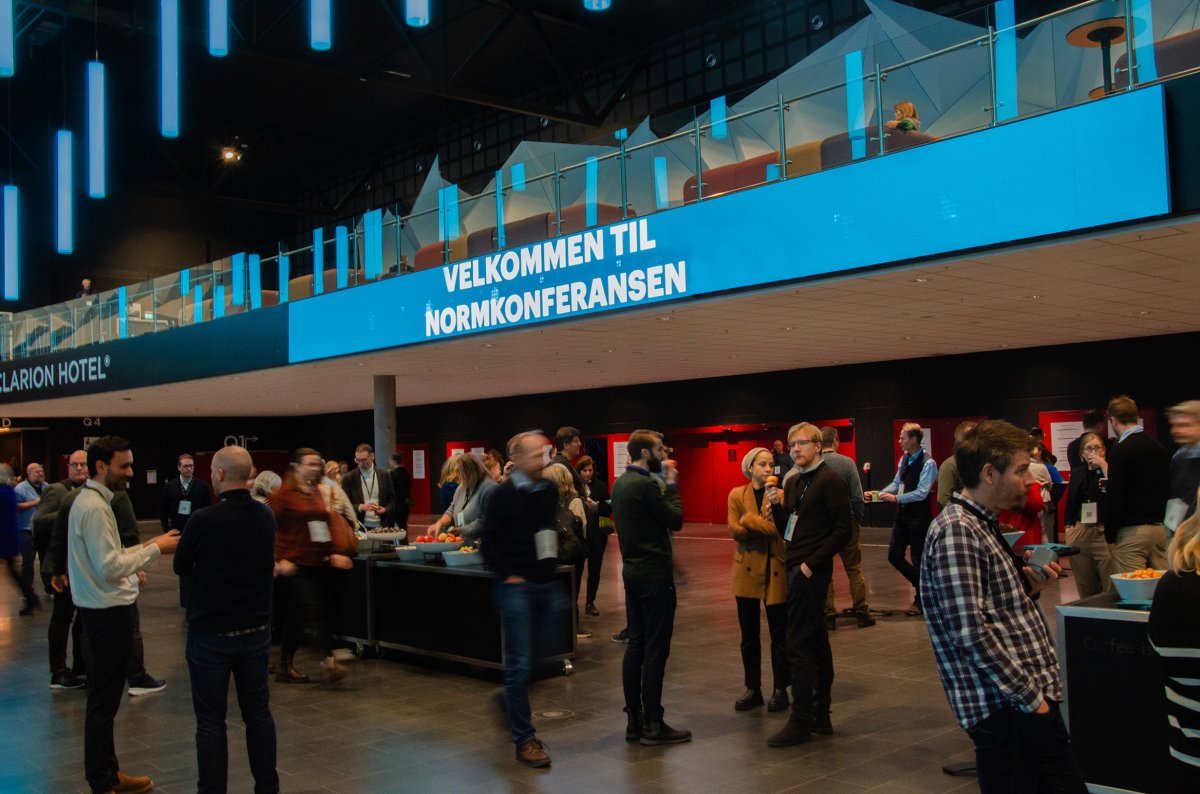 Mange mennesker som passerer forbi eller snakker sammen unde ret skilt med teksten «Velkommen til Normkonferansen»