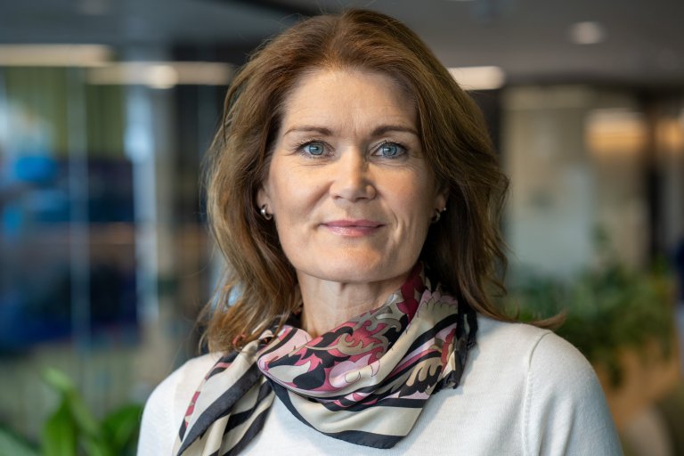 bilde av kvinne med hvit genser og skjerf i et kontorlandskap med grønne planter i bakgrunnen