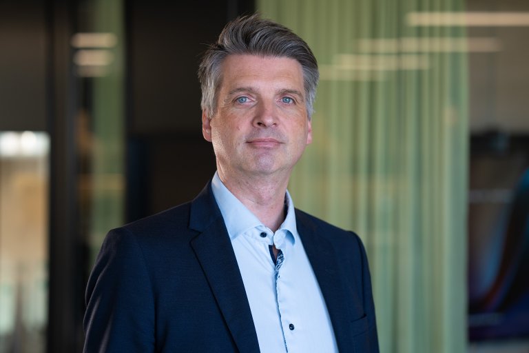 Mann med dressjakke og blå skjorte står foran kontorlandskap og grønn gardin